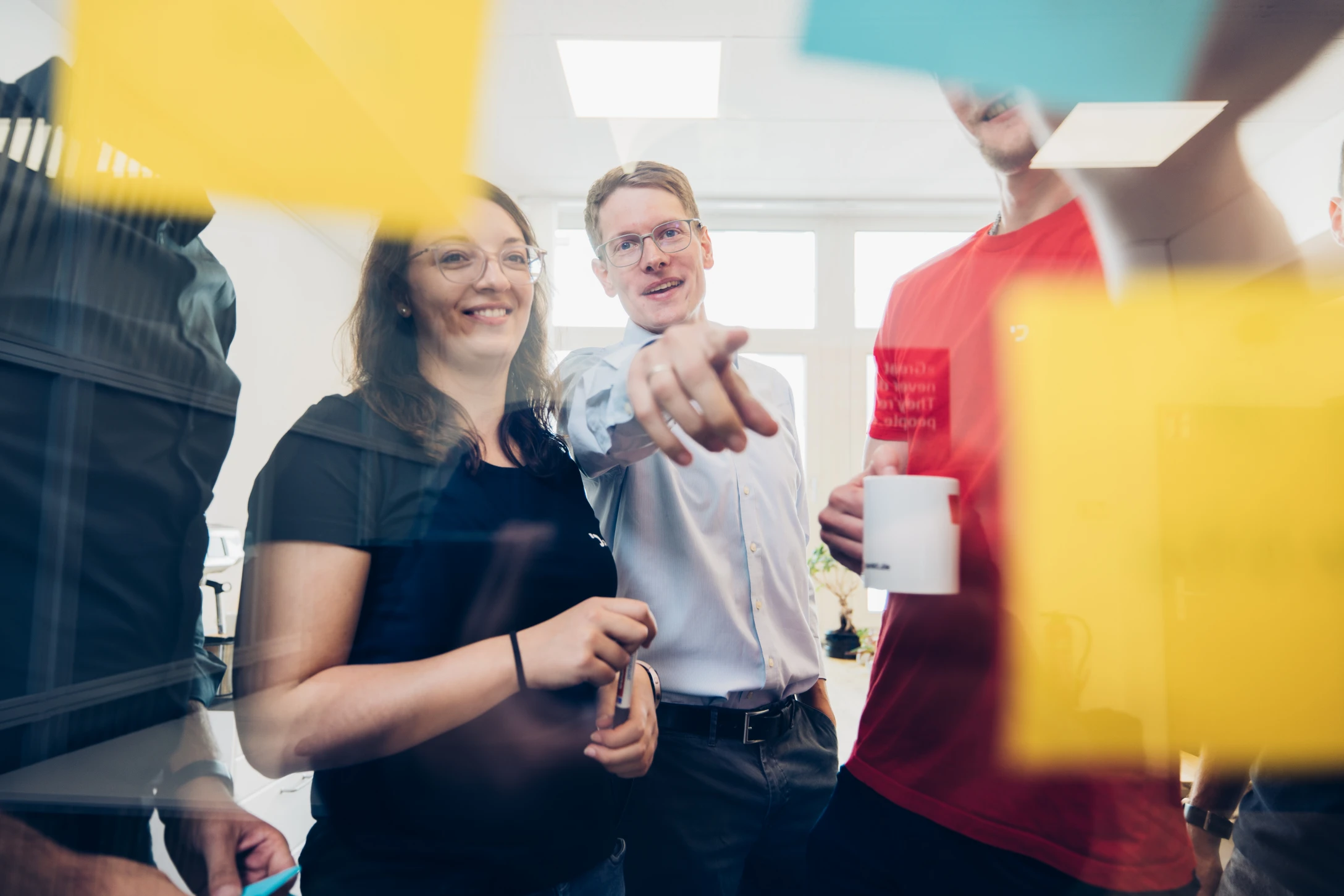 Ein Team von Mitarbeitenden steht vor einem transparenten Whiteboard, das mit bunten Haftnotizen bedeckt ist, und sammelt Ideen..