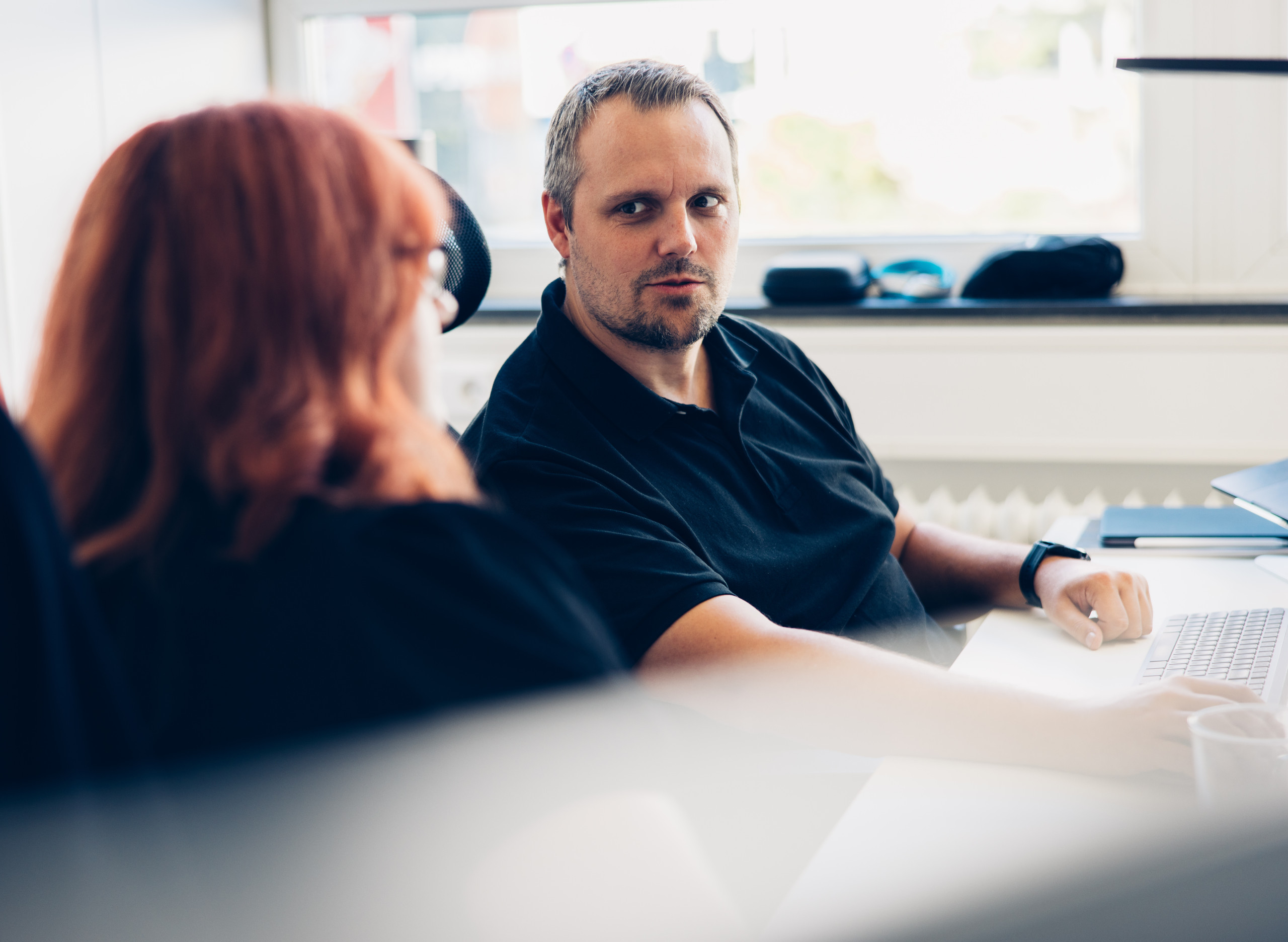 Ein Mann sitzt in einem Büro an einem Schreibtisch und blickt aufmerksam zu einer Kollegin