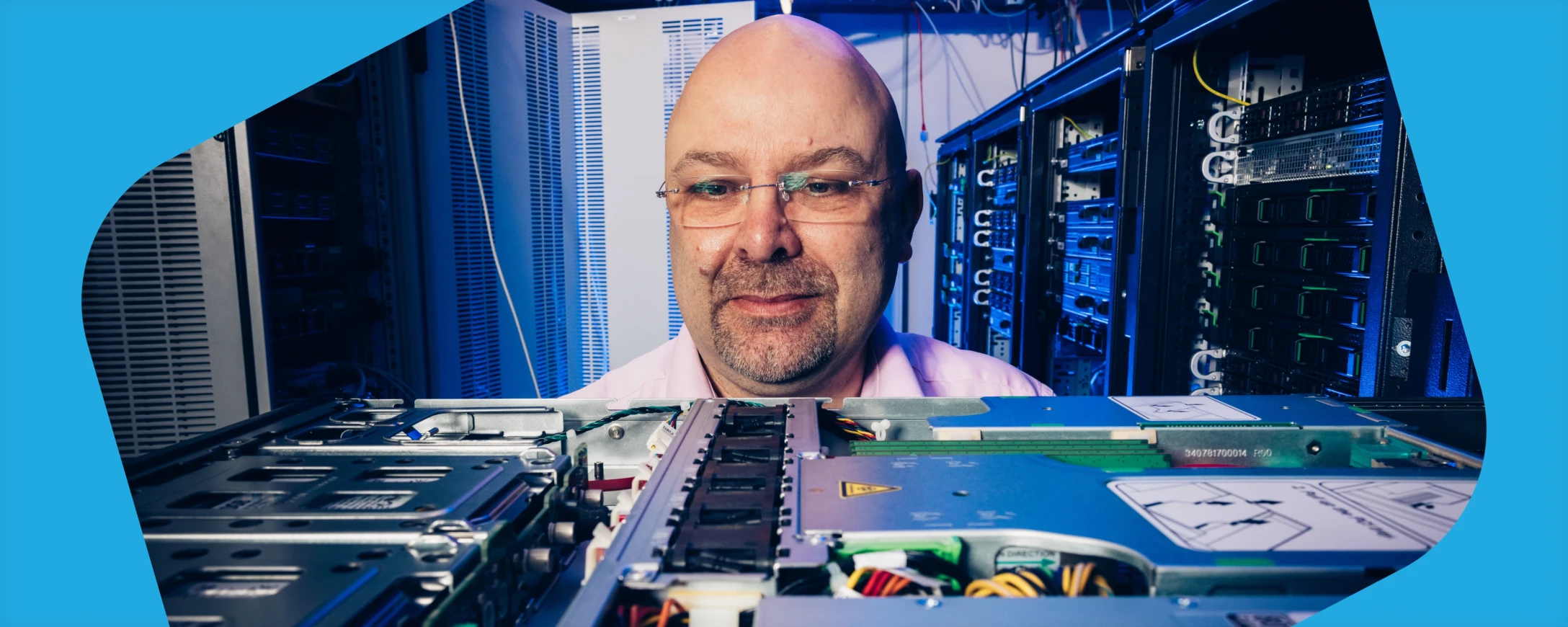 Ein Mann mit Bart und Brille schaut lächelnd auf ein Servermainboard. Im Hintergrund sieht man einen Serverschrank.