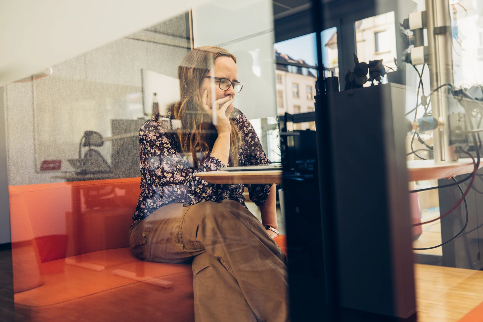 A young woman with glasses sits thoughtfully in front of a computer in a modern office.